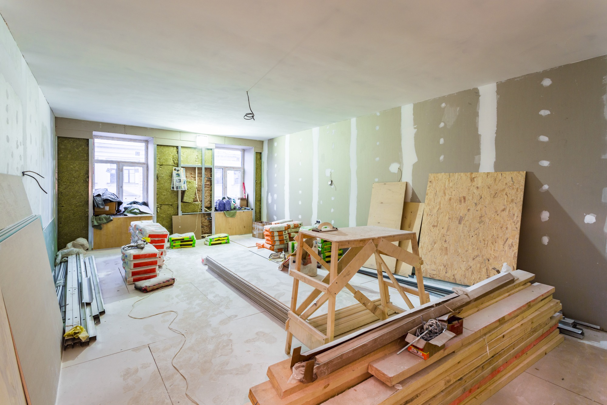 Renovation in progress: a bright room with bare walls, wood panels, and construction materials scattered throughout. Tools and supplies are visible.