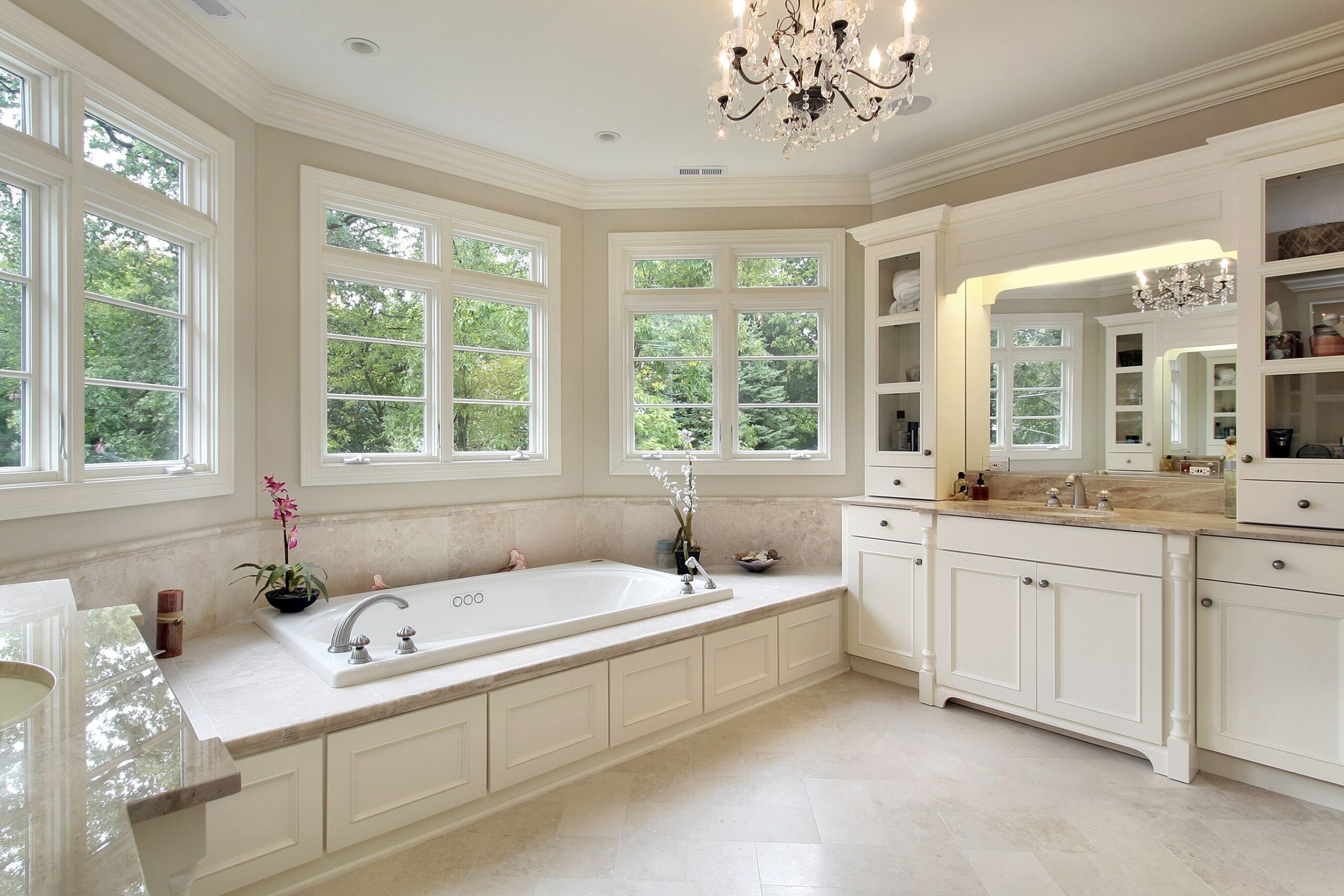 Bright, elegant bathroom featuring a freestanding tub, double sink vanity, and multiple windows with a view of greenery. A chandelier adds a touch of luxury.