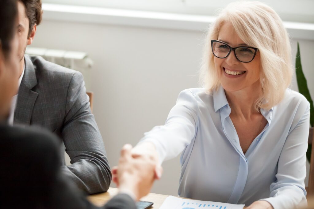 A confident woman with glasses smiles and shakes hands during a business meeting, signifying a successful partnership or agreement.