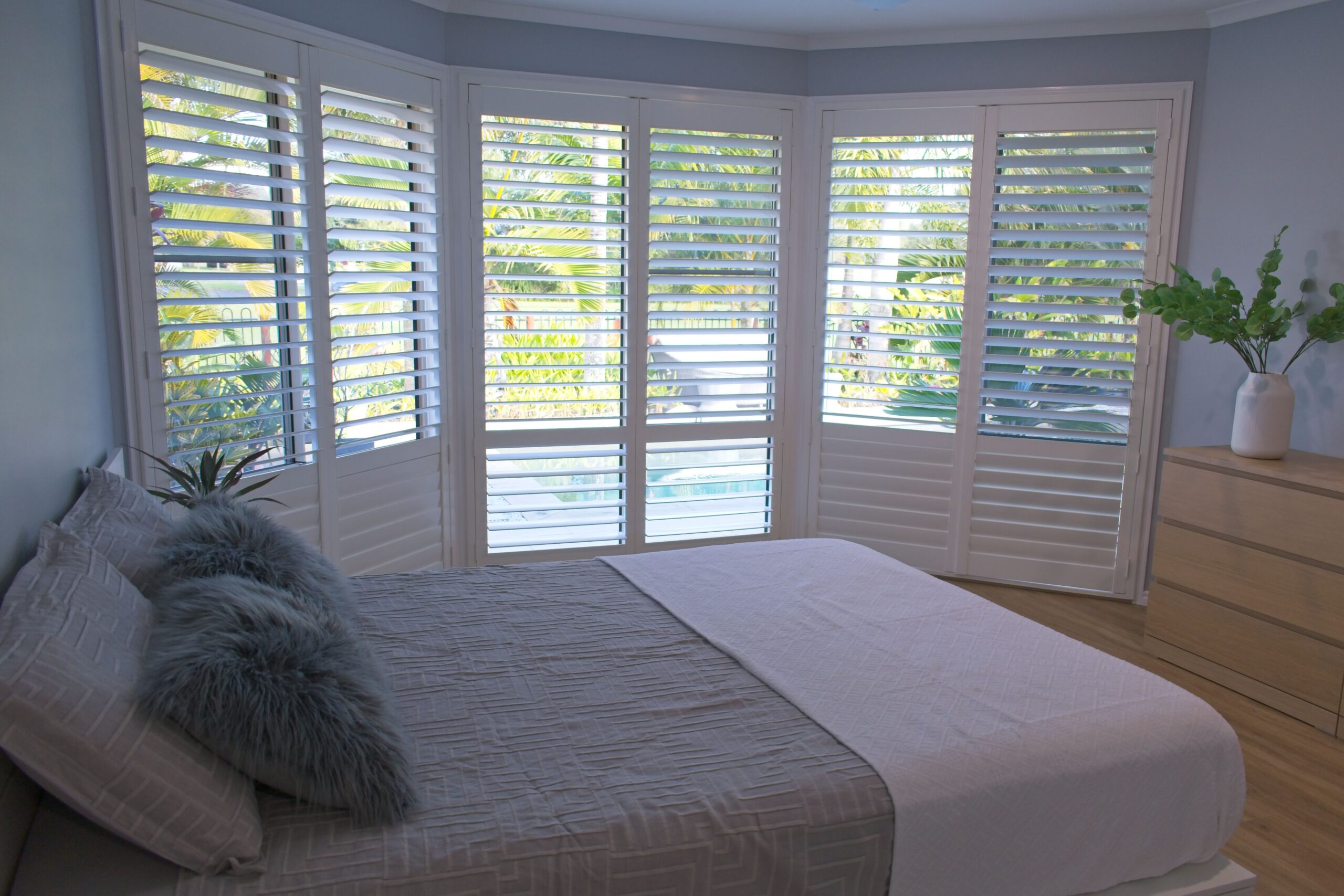 Cozy bedroom featuring a light-colored bed with fluffy pillows, large shuttered windows, and green plants, creating a bright and inviting atmosphere.