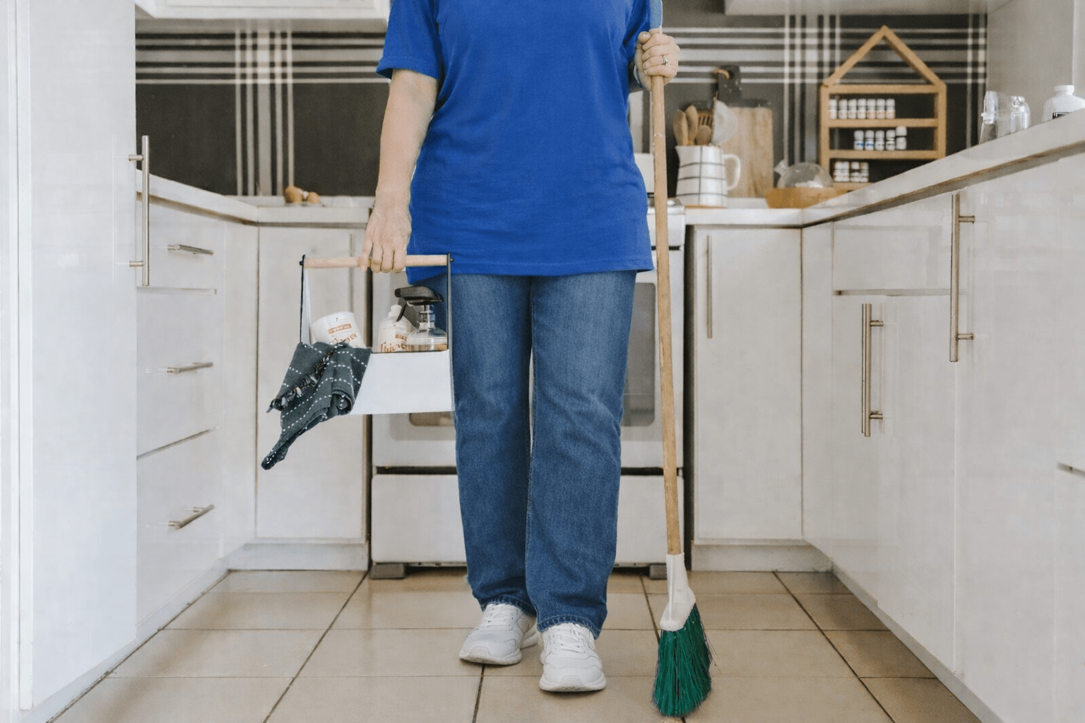A person in a blue shirt holds a cleaning caddy filled with supplies in one hand and a broom in the other, standing in a clean kitchen.