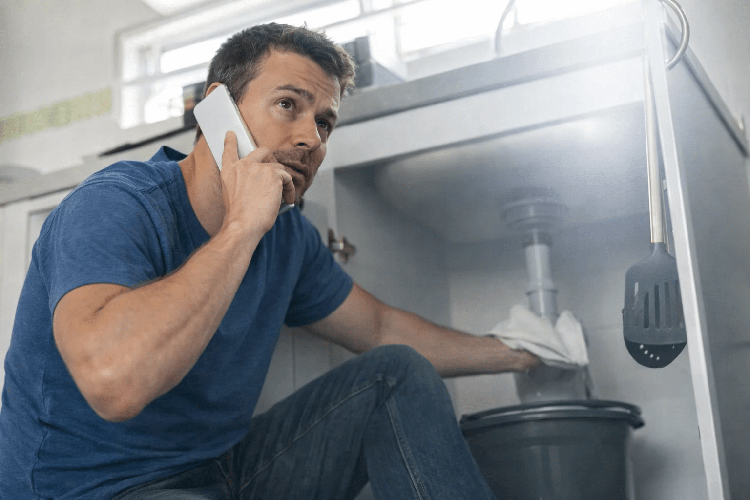 Man on the phone crouching under a kitchen sink, holding a towel, appearing to troubleshoot a plumbing issue. Relevant for home repair topics.